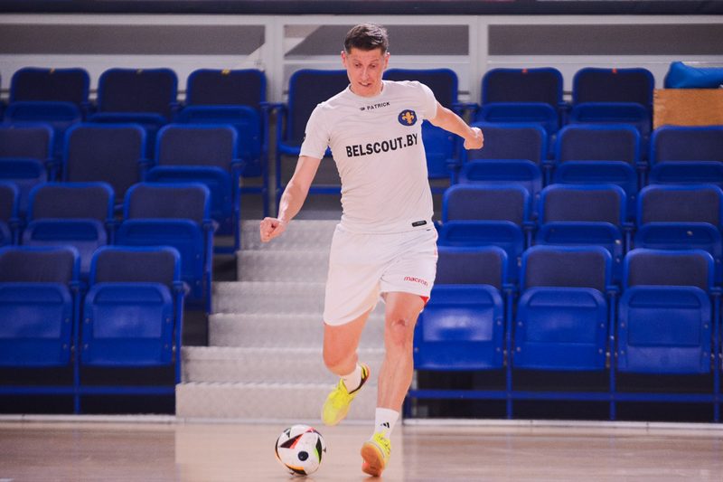 A futsal player in a white BELSCOUT.BY shirt carrying the ball across an indoor court with rows of blue seats behind him.