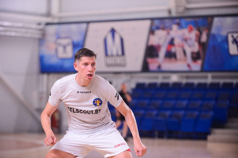 A futsal player in a white BELSCOUT.BY shirt braced low to press an opponent on an indoor court.