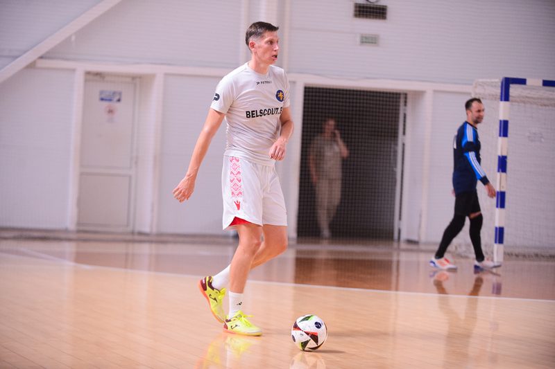 A futsal player in a white kit taking a ball down on an indoor court while an opponent watches from the edge of the frame.