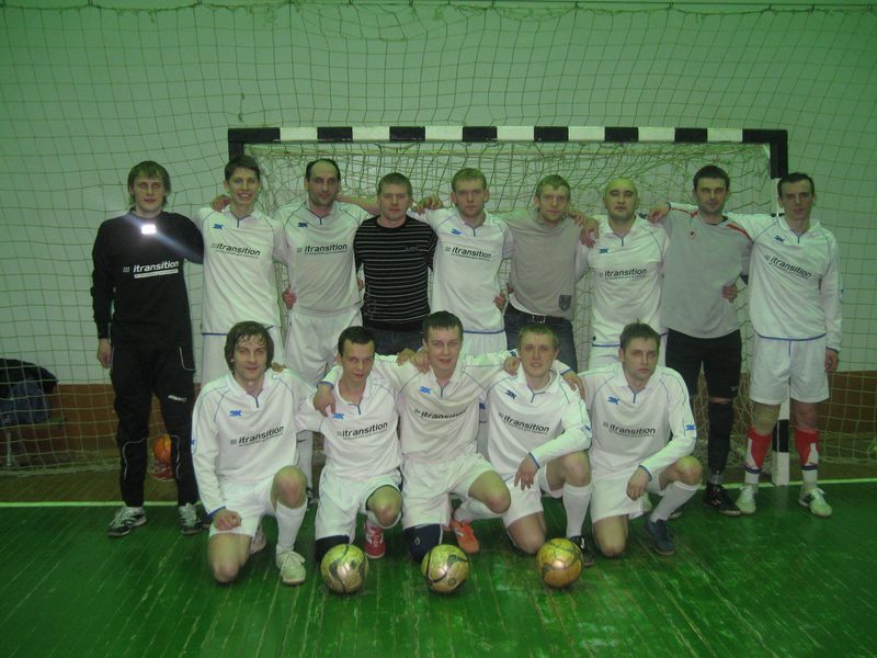 A futsal squad in white kit lined up with their coach in front of a green goal in a Minsk sports hall.