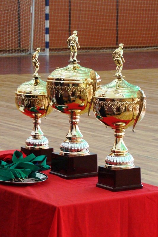 Three gold futsal trophies standing on a red presentation table in a sports hall.