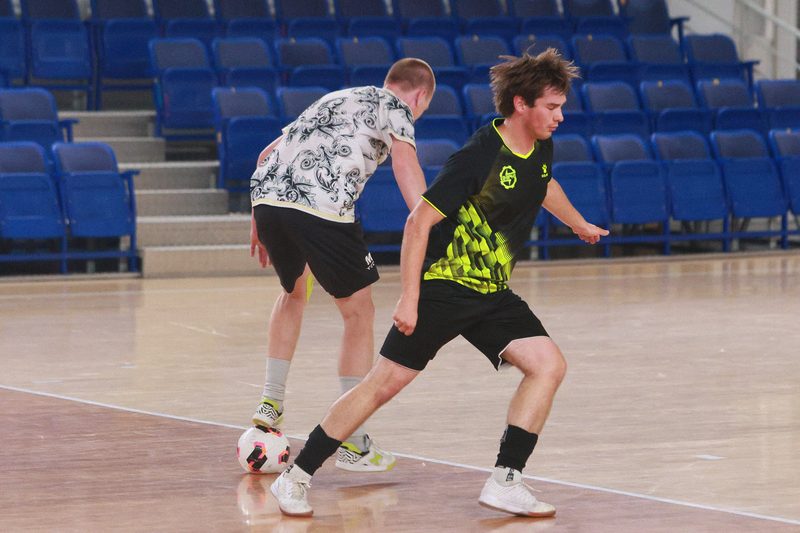 Two futsal players, one in white and one in black and neon, contesting the ball in front of empty blue seats.