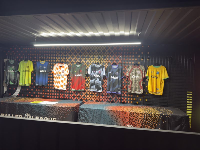 Numbered football shirts hanging in wooden lockers under a lit Baller League sign.