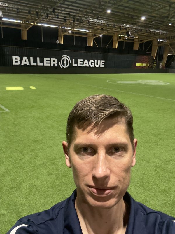 Anton Abyzov pitchside at the Baller League Miami arena with the league boards lit behind him.