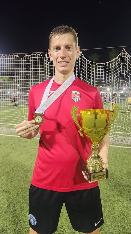 Anton Abyzov in a red Patagonia FC shirt holding the gold cup in one hand and a winner's medal in the other.