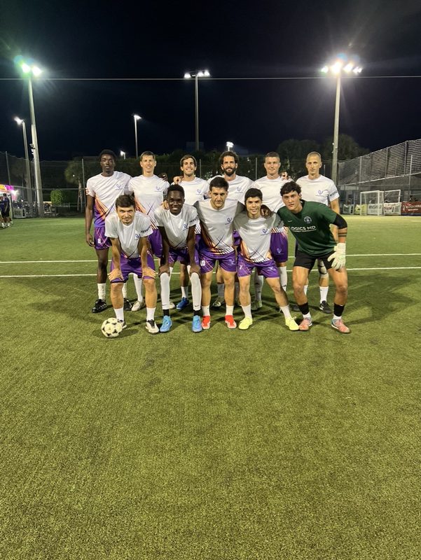 An eight a side squad in white shirts and purple shorts lined up on a floodlit pitch in Miami.
