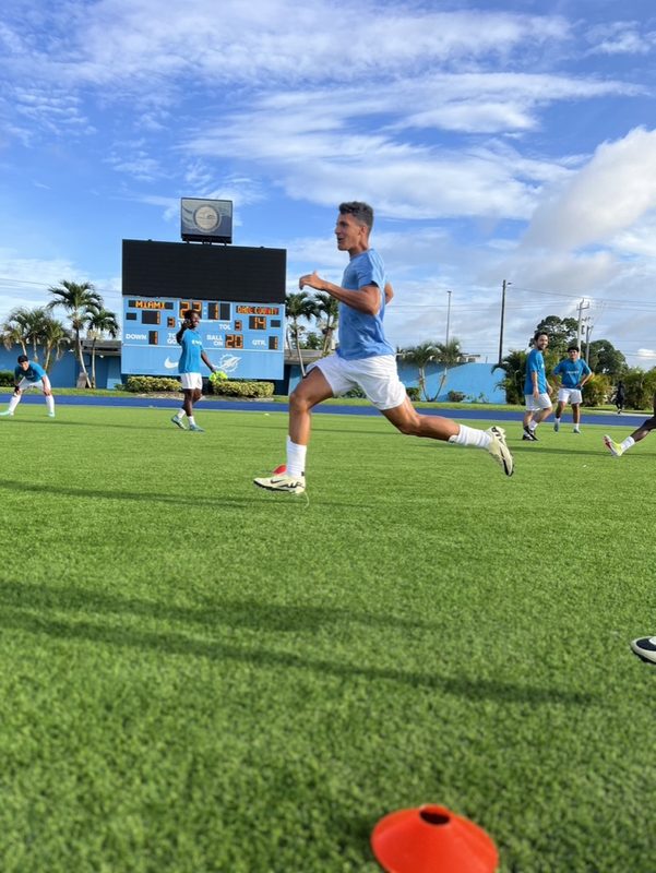 Anton Abyzov at full stride on the turf in a light blue kit, scoreboard behind him.