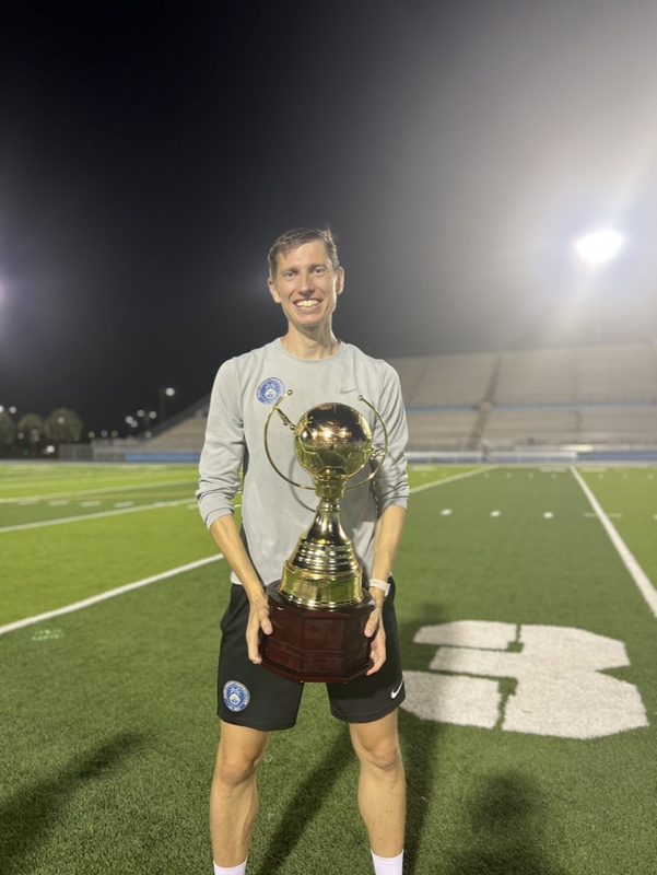 Anton Abyzov holding the USSL trophy at waist height on the halfway line under floodlights.