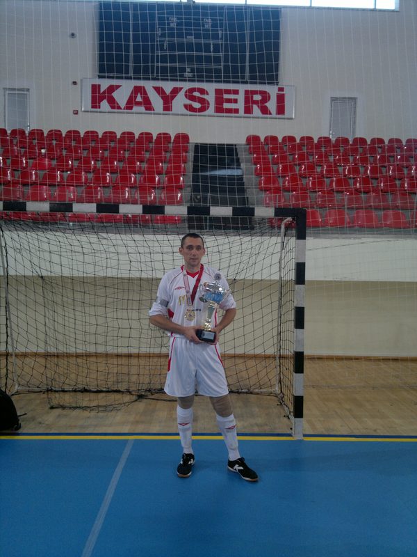 A player in the white Belarus shirt holding the cup in front of the goal, with the red Kayseri banner across the wall behind.