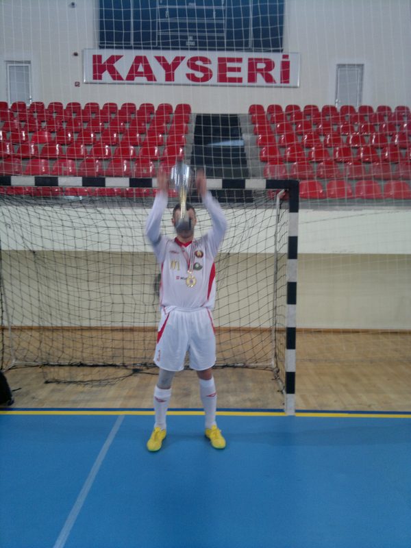 A player in the white Belarus shirt lifting the championship cup above his head in the goalmouth under the Kayseri banner.