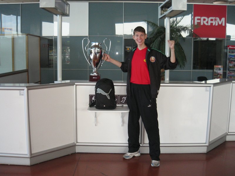 Anton Abyzov in a Belarus tracksuit standing beside the European Championship cup on an airport counter in Nantes, one arm raised.