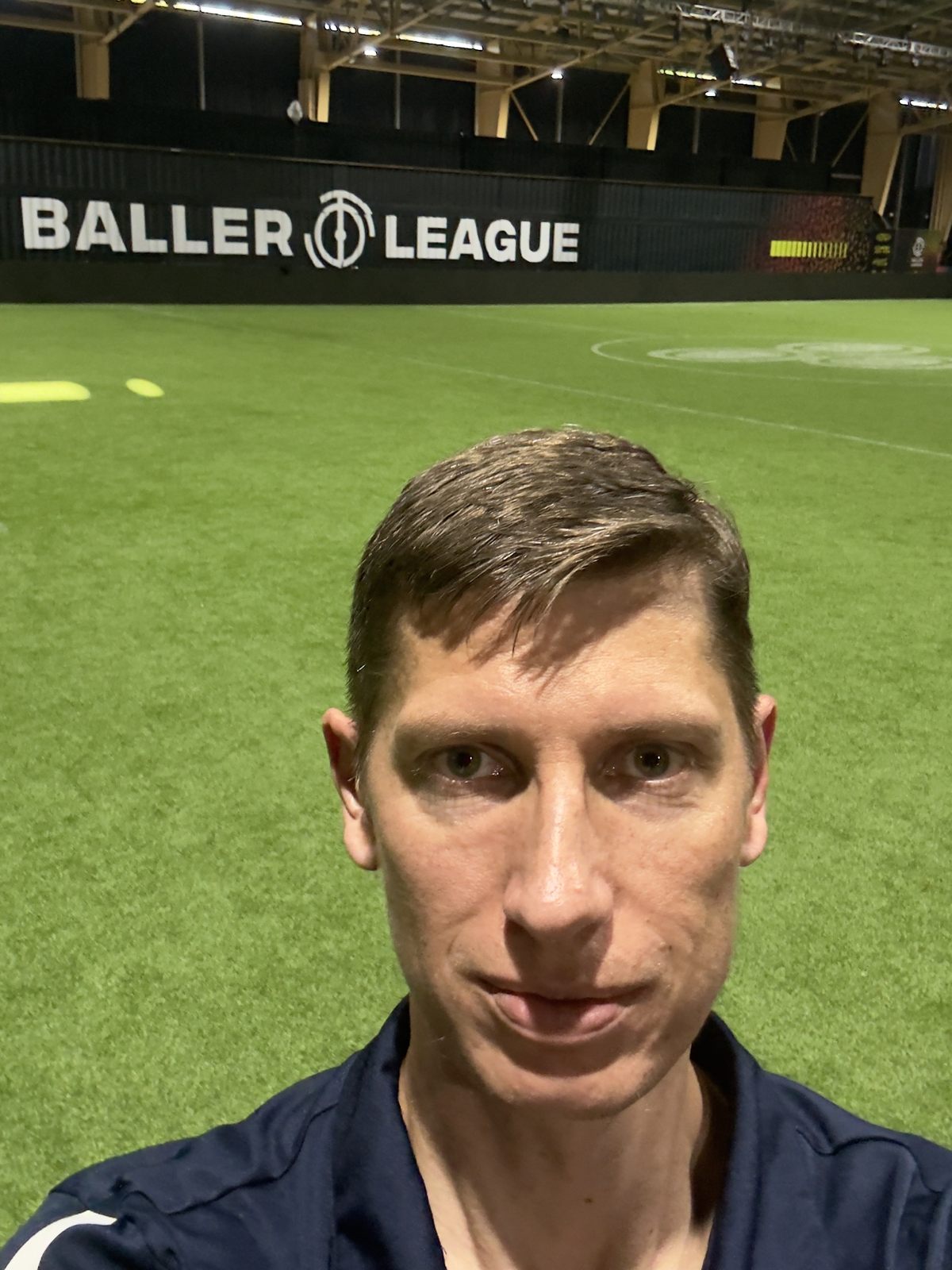 Anton Abyzov pitchside at the Baller League USA arena in Miami, league boards and floodlights behind him.