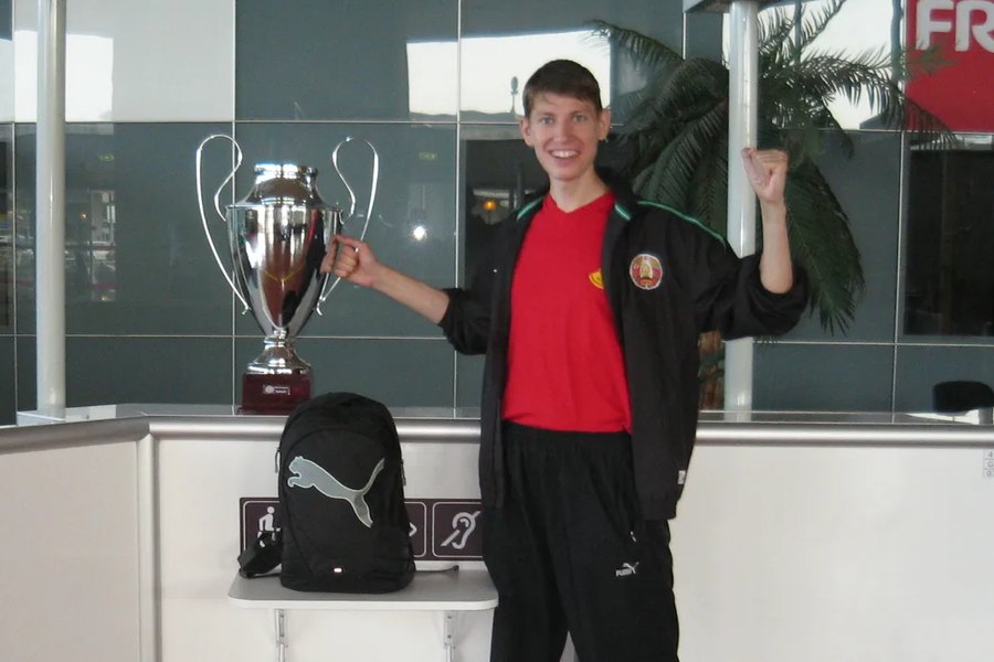 Anton Abyzov in a Belarus tracksuit beside the European Championship cup on an airport counter in Nantes, one arm raised.