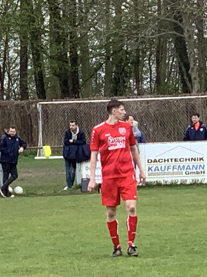 Anton Abyzov standing in the red SV Rangsdorf 28 kit in front of the home dugout at Stadion Rangsdorfer See.