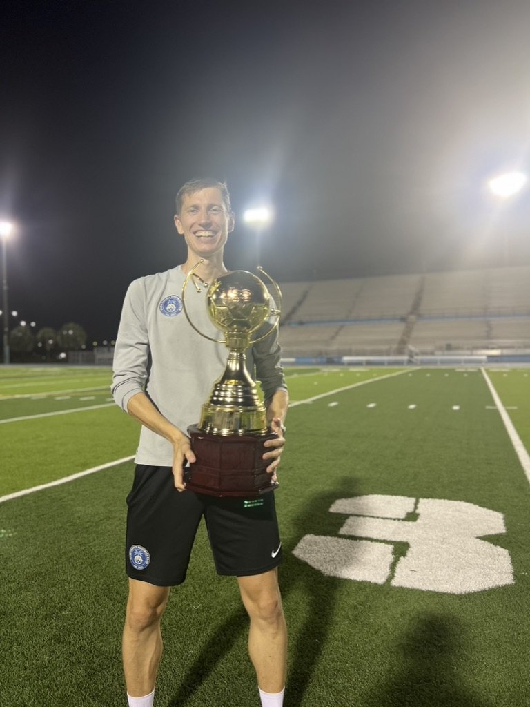 Anton Abyzov holding a trophy on a floodlit pitch in Miami.